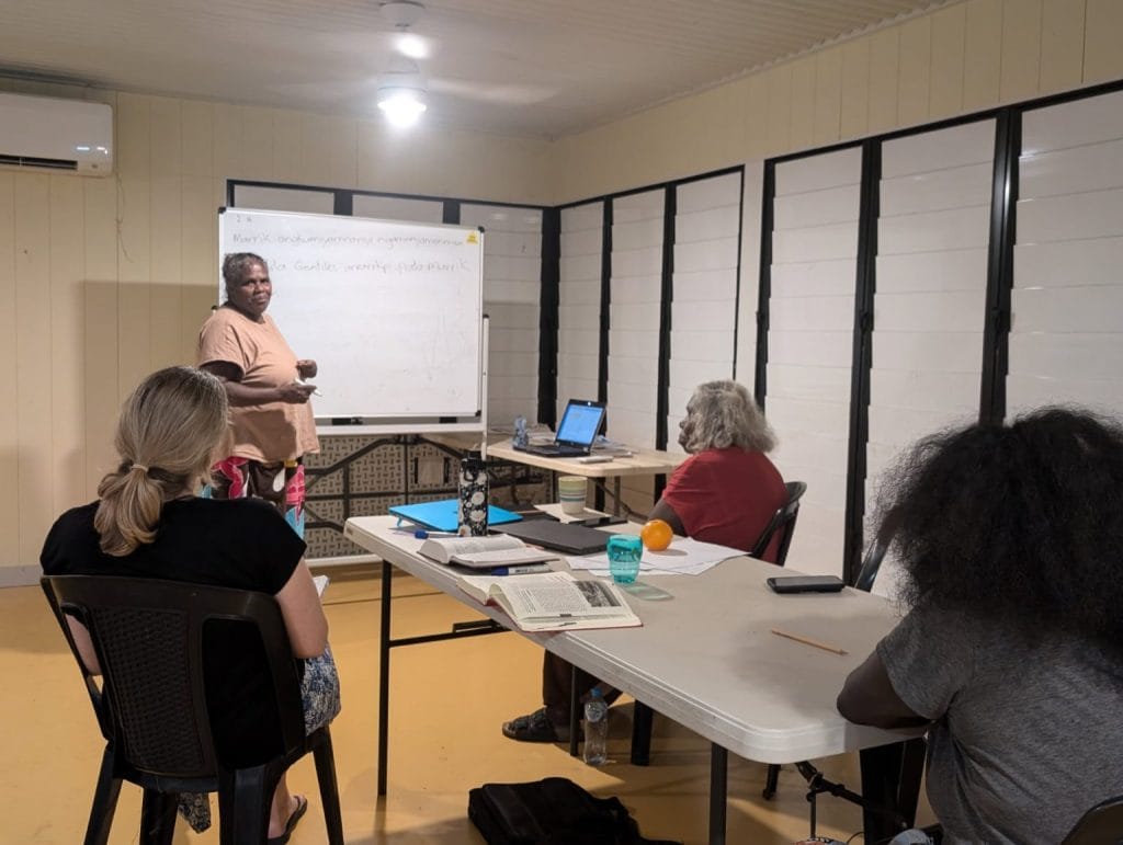 Anne (seated, facing away from camera) listening to a whiteboard presentation from an indigenous Australian.